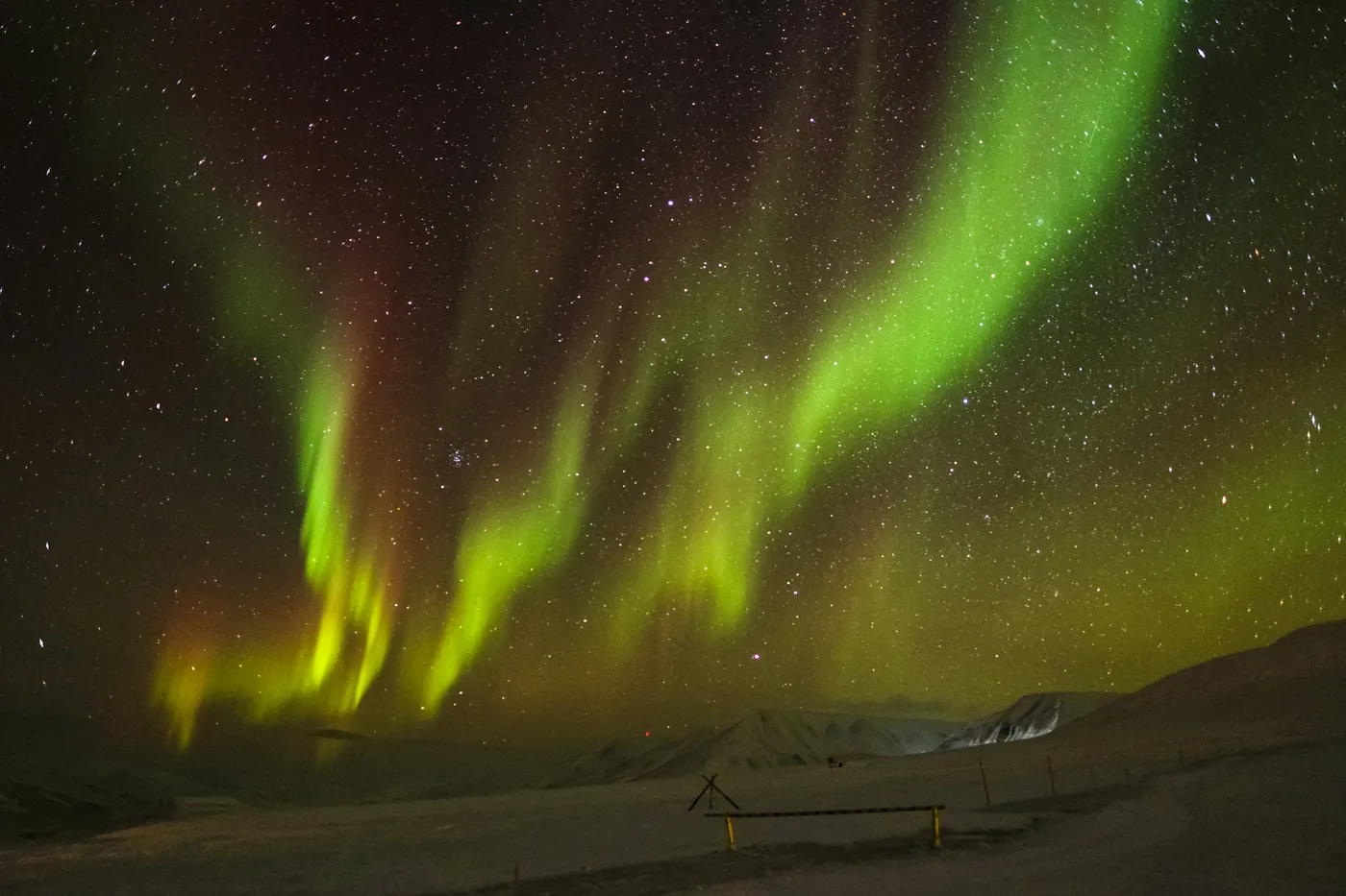 Grüne und gelbe Polarlichter wölben sich unter einem Sternenhimmel über ein verschneites Tal