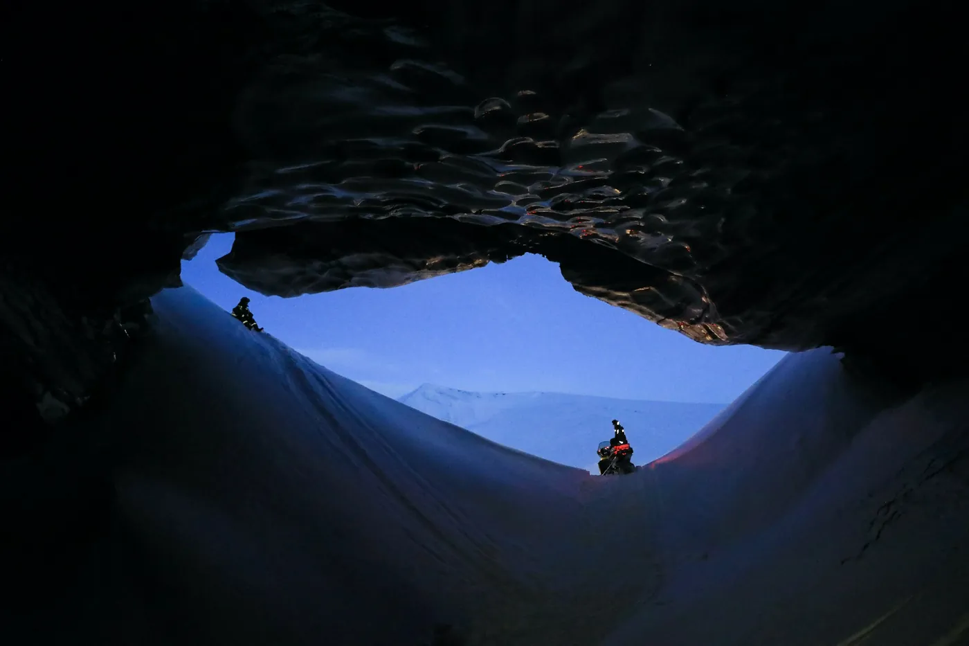 Blick aus einer dunklen Eishöhle auf einen verschneiten Hang mit zwei Personen und einem Schneemobil