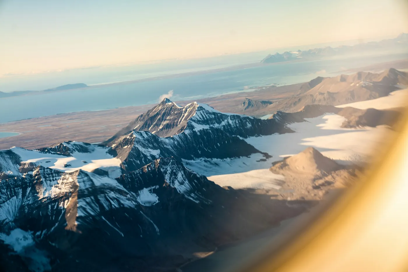Cime innevate e ghiacciai visti dal finestrino di un aereo, con un fiordo in lontananza